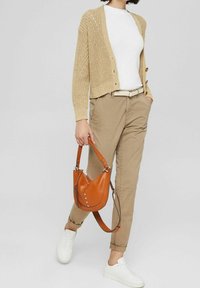 Vrouw draagt een beige vest, wit T-shirt, kaki broek en witte sneakers, en houdt een bruine leren handtas vast aan de handvatten.