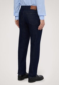 Jeans de mezclilla azul oscuro con un corte recto, que cuentan con un parche de cuero marrón en la cinturilla y en los bolsillos traseros. Combinados con zapatos negros.