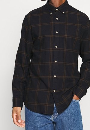 Mand iført en mørkebrun ternet knap-op skjorte med lange ærmer og blå jeans, den ene hånd i lommen, foran en hvid baggrund.