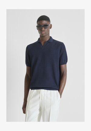 Muškarac u mornarskoplavoj kratkoročavnoj pletenoj polo majici i bijelim plisiranim hlačama, pozira s jednom rukom u džepu i nosi velike crne sunčane naočale.