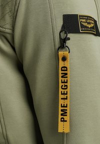 Maslinasto zelena jakna s rombićastim ramenom, koja sadrži crni pečat s natpisom "PME Legend" u žutoj boji i žutu ručicu za zatvarač s natpisom.