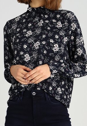 Femme portant un chemisier noir à manches longues avec un motif floral blanc et un jean foncé, debout les mains librement jointes à la taille.