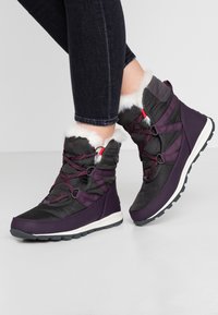 Lila und schwarze isolierte Stiefel mit Kunstfellfutter, ausgestattet mit einer strukturierten Oberfläche, einem Schnürsystem und einer weißen Zwischensohle auf einer gummierten Außensohle.