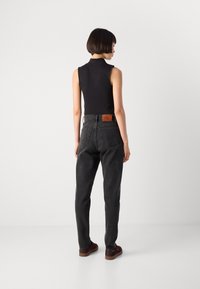 Moteris su trumpais plaukais stovi atsisukusi atgal, vilkinti be_rankovių juodą marškinėlį, aukšto liemens tamsiai pilkas džinsus ir tamsiai raudonus sportinius batelius ant baltos fono.