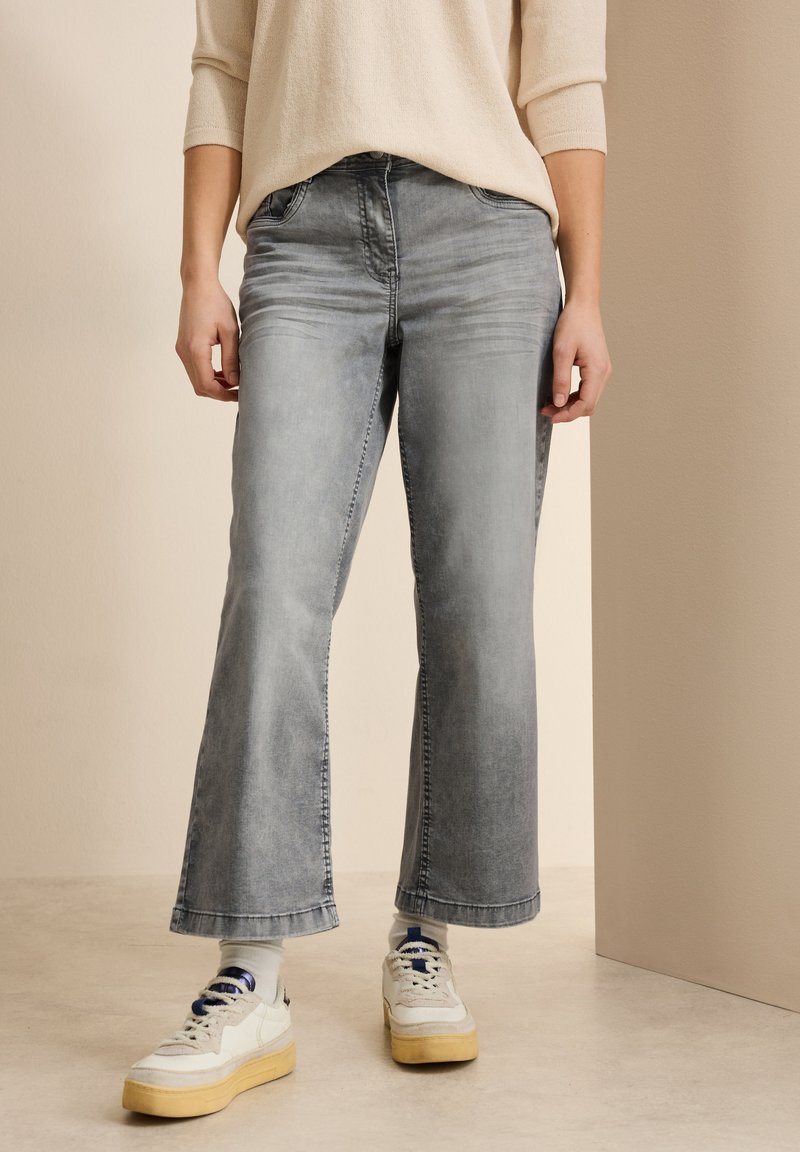 Jean large gris avec une finition délavée, présentant un design classique à cinq poches. Associé à des baskets blanches et des chaussettes à motifs.