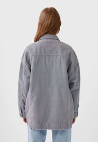 Persoon met lang lichtbruin haar, gekleed in een oversized grijs corduroy jack en lichtblauwe jeans, met de rug naar de camera gericht tegen een effen achtergrond.