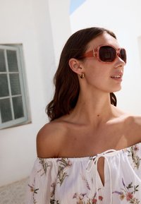 Solglasögon med bruna glas och en tjock, transparent orange båge. Designlogotypen syns på skalmarna. Modellen bär en blommig off-shoulder-topp.