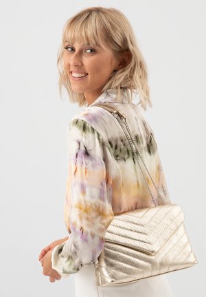 Metalen gouden schoudertas met een chevronpatroon, leren textuur en kettingriem, gedragen over een multicolored blouse.
