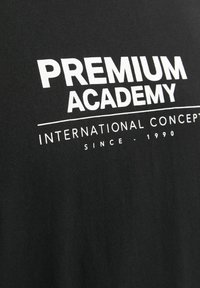 Črna bombažna majica s belim tiskom: "PREMIUM ACADEMY INTERNATIONAL CONCEPTS SINCE - 1990," moderna pisava s horizontalno poravnavo.