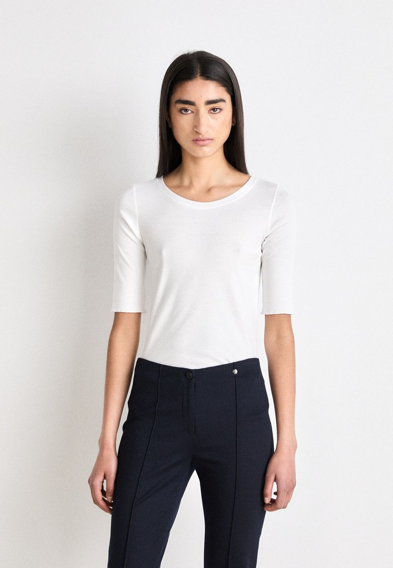 Top bianco aderente a maniche corte con scollatura rotonda, abbinato a pantaloni sartoriali blu scuro con chiusura a bottone e dettagli delle cuciture verticali.