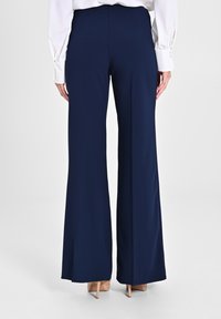Pantalons larges de couleur marine en tissu lisse, dotés d'une taille haute, de coutures sur les côtés et d'une silhouette fluide. Associés à des escarpins nus.