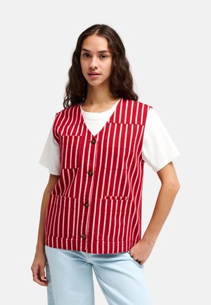Junge Frau mit langen dunklen Haaren, die ein weißes kurzärmeliges Hemd und eine rot gestreifte Weste mit Knöpfen und Taschen trägt, kombiniert mit hellblauen Jeans.