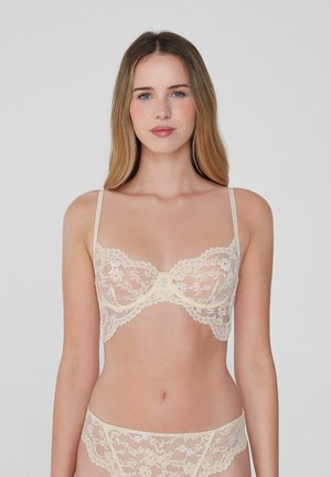 Koronkowy biustonosz typu bralette i pasujące figi w delikatnym beżu. Regulowane ramiączka, falbaniasty brzeg, kwiatowy wzór, półprzezroczysty materiał, wsparcie w postaci fiszbiny.