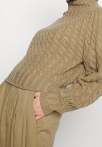 Pull en maille torsadée beige avec un col montant, présentant des motifs texturés et des manches amples, associé à un pantalon taille haute beige clair.
