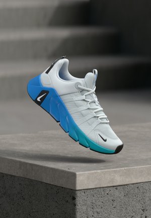 Baskets athlétiques gris clair et turquoise avec logos Nike noirs et blancs, flottant au-dessus d'un rebord en béton sur un fond gris flou.