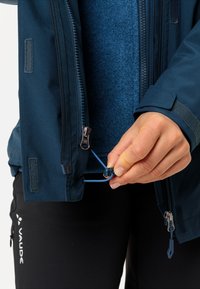 Giacca impermeabile blu navy con zip frontale, cordino regolabile e polsini in velcro. Abbinata a pantaloni neri con logo.