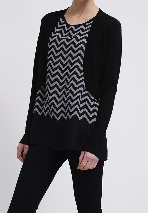 Personne portant un cardigan noir ouvert sur un haut à motif chevron noir et gris, accompagné d'un pantalon noir, sur un fond uni.