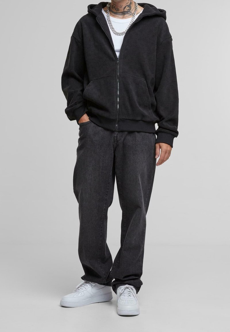 Homme portant un sweat à capuche noir avec fermeture éclair, un jean ample foncé, des baskets blanches, un débardeur blanc, des chaînes argentées et des tatouages visibles au cou.