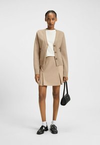 Beżowy kardigan prążkowany z guzikami z przodu i kieszeniami, zestawiony z dopasowaną beżową spódnicą w prążki z plisami. Czarne buty i torebka.