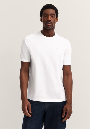 Mann in einem schlichten weißen Kurzarm-T-Shirt und dunkelblauen Jeans, der vor einem schlichten hellen Hintergrund steht.