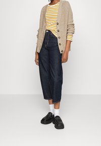 Beige stickad cardigan med knappar, gul och vit randig skjorta, mörkblå avklippta jeans och svarta plattformsskor med vita strumpor.