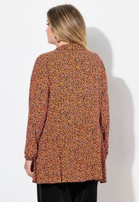 Túnica de manga larga en color naranja con un patrón de leopardo en negro, morado y rojo. Cuenta con un corte recto y un detalle de pliegues en la parte posterior.
