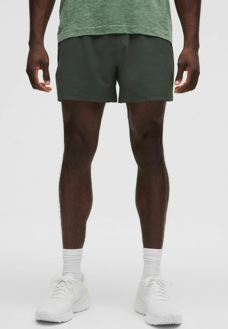 Mörkgröna sportshorts i lätt material, tillsammans med vita sneakers och ankelsockor, visade på en neutral bakgrund.