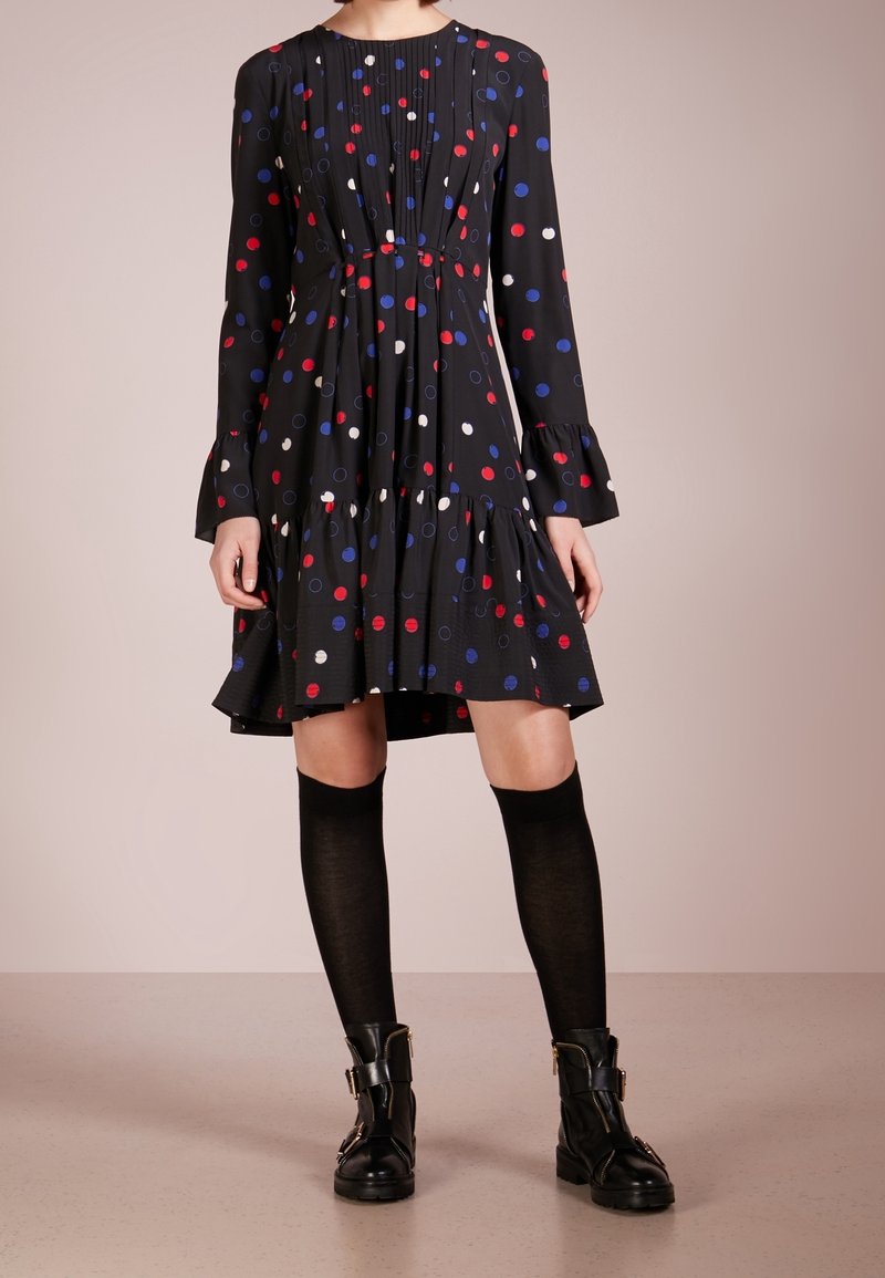 Svart långärmad klänning med röda, blåa och vita prickar, plisserad bodice, volangkant, ihopparad med knähöga svarta strumpor och svarta stövlar.