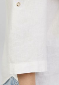 Chemise en lin blanc avec des manches retroussées, présentant une texture subtile et un accent de bouton rond et transparent près du poignet.