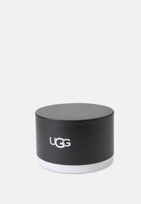 Ottomán de almacenamiento redondo negro hecho de cuero sintético con una textura suave, presenta una base blanca y el logo de UGG impreso en blanco.