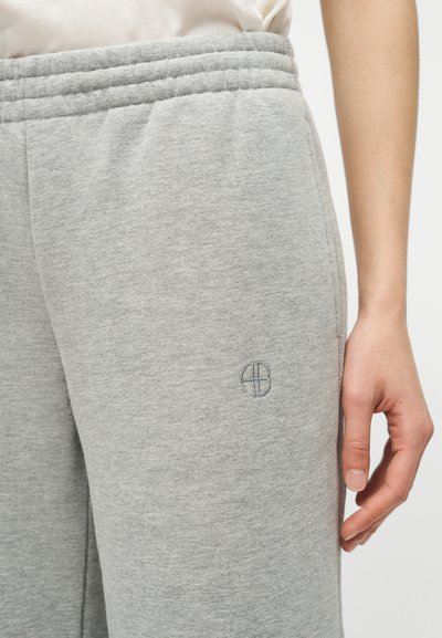 Gros plan sur une personne portant un pantalon de survêtement gris clair avec une taille élastique et un petit logo circulaire brodé près de la hanche.