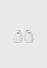 Adidași din piele albă cu o textură netedă, profil jos și talpă din cauciuc. Partea din spate are un logo negru, design minimalist și linii curate.