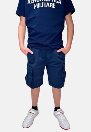 T-shirt in cotone blu navy con testo "AERONAUTICA MILITARE"; pantaloni cargo blu navy con tasche laterali; sneakers bianche con accenti rossi.