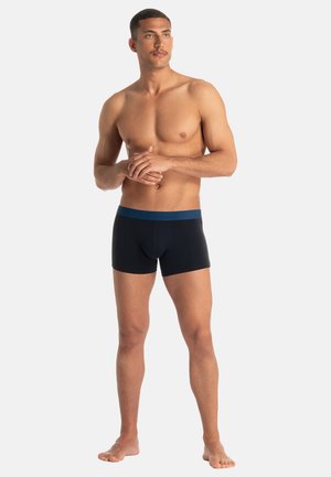 Schwarze Boxershorts mit einem blauen Taillenbund, gefertigt aus einem glatten, dehnbaren Material. Das Modell steht barfuß und präsentiert Passform und Design.