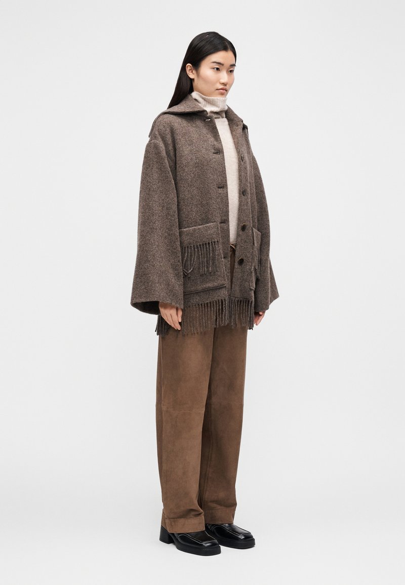 Brun uldfrakke med hætte, knaplukning, forlommer med frynser, båret over en lys rullekrave og brune ruskindsbukser, kombineret med sorte sko.