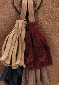 Tasseles en tres colores—beige, burdeos y azul oscuro—hechos de suave ante, sujetos con un lazo a un fondo texturizado marrón.