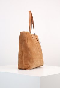 Tan suède tote bag met leren handvatten staat rechtop op een witte pedestal tegen een effen achtergrond.