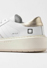 Zapatilla blanca de cuero con un acento en el talón de metal dorado, suela de plataforma elevada y detalle del logo cuadrado negro en el lateral. Plantilla texturizada en gris.