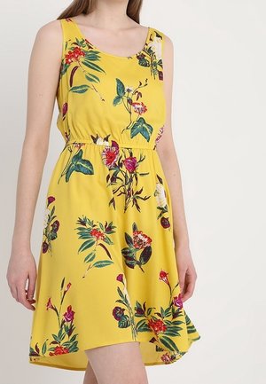 Femme portant une robe jaune sans manches à imprimé floral coloré et taille cintrée, sur un fond uni.
