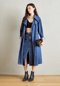 Long manteau en denim avec une fente profonde, porté sur un crop top noir et une jupe en denim taille haute. Présente des manches retroussées et des bottines noires.