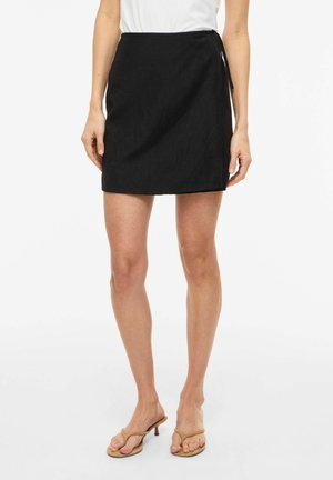 Femme portant une mini-jupe portefeuille noire avec un lien sur le côté, un haut blanc et des sandales beiges à talons, debout sur un fond blanc.