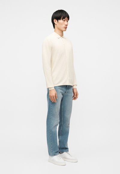 Homme debout portant une chemise crème à manches longues boutonnée, un jean bleu et des baskets blanches, sur un fond blanc uni.