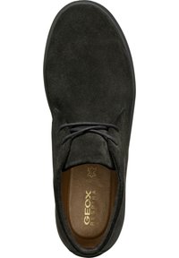 Zapato de ante negro con puntera redonda, que presenta cordones oscuros, una plantilla acolchada y una suela de goma. Logo de la marca en dorado visible en el interior.