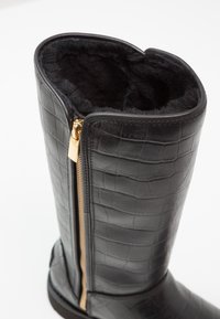 Botte noire montant jusqu'au genou avec texture crocodile, fermeture éclair dorée sur le côté et doublure intérieure en fourrure noire douce dans la tige.