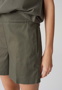 Olivově zelené šortky vyrobené z bavlněného směsového materiálu, s uvolněným střihem, bočními kapsami a prošitým lemem. Hladká textura s minimálními detaily.