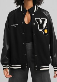 Chaqueta varsity negra con rayas blancas, que presenta una gran letra 'V' bordada y un diseño de flores. Elaborada en lana y cuero sintético.