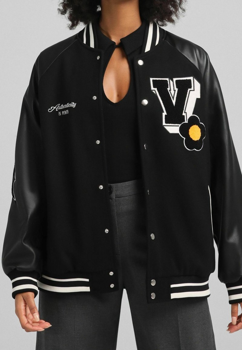 Chaqueta varsity negra con rayas blancas, que presenta una gran letra 'V' bordada y un diseño de flores. Elaborada en lana y cuero sintético.