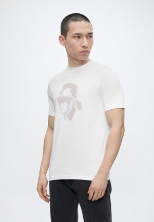 Νέος άνδρας με κοντά μαλλιά που φοράει λευκό κοντομάνικο μπλουζάκι με γραφικό πρόσωπο στιλιζαρισμένο και μαύρο παντελόνι, κοιτάζοντας προς το πλάι.