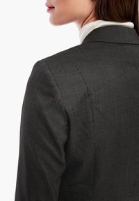 Czarny żakiet o teksturowanej powierzchni z subtelnym geometrycznym wzorem, dopasowany kształt i usztywnione ramiona, połączony z białym golfem pod spodem.