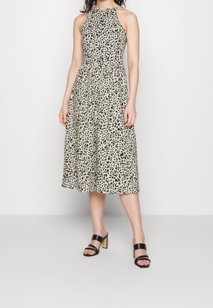 Kobieta w bezowej sukience midi bez rękawów z czarnymi nieregularnymi plamami oraz czarnych sandałach na obcasie z odkrytymi palcami na jednolitym białym tle.